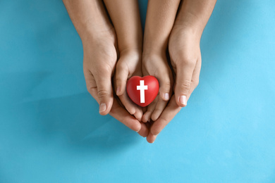 Image of Mother and her child holding heart with cross symbol on light blue background, top view. Christian religion