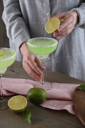 Photo of Woman squeezing lime juice into glass with delicious Margarita cocktail at wooden table, closeup