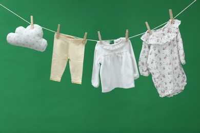 Photo of Different baby clothes and cloud shaped pillow drying on laundry line against green background