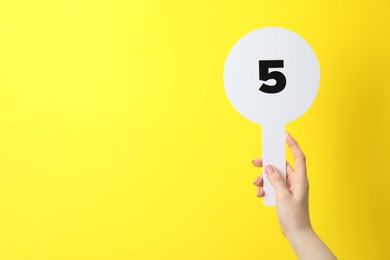 Photo of Woman holding auction paddle with number 5 on yellow background, closeup. Space for text