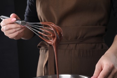Photo of Woman holding whisk with flowing chocolate cream on black background, closeup