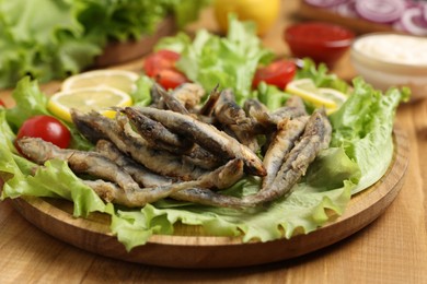 Photo of Delicious fried anchovies served with lemon, tomatoes and lettuce leaves on wooden table, closeup