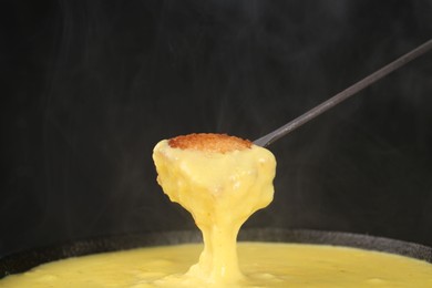 Dipping piece of bread into fondue pot with tasty melted cheese against dark gray background, closeup