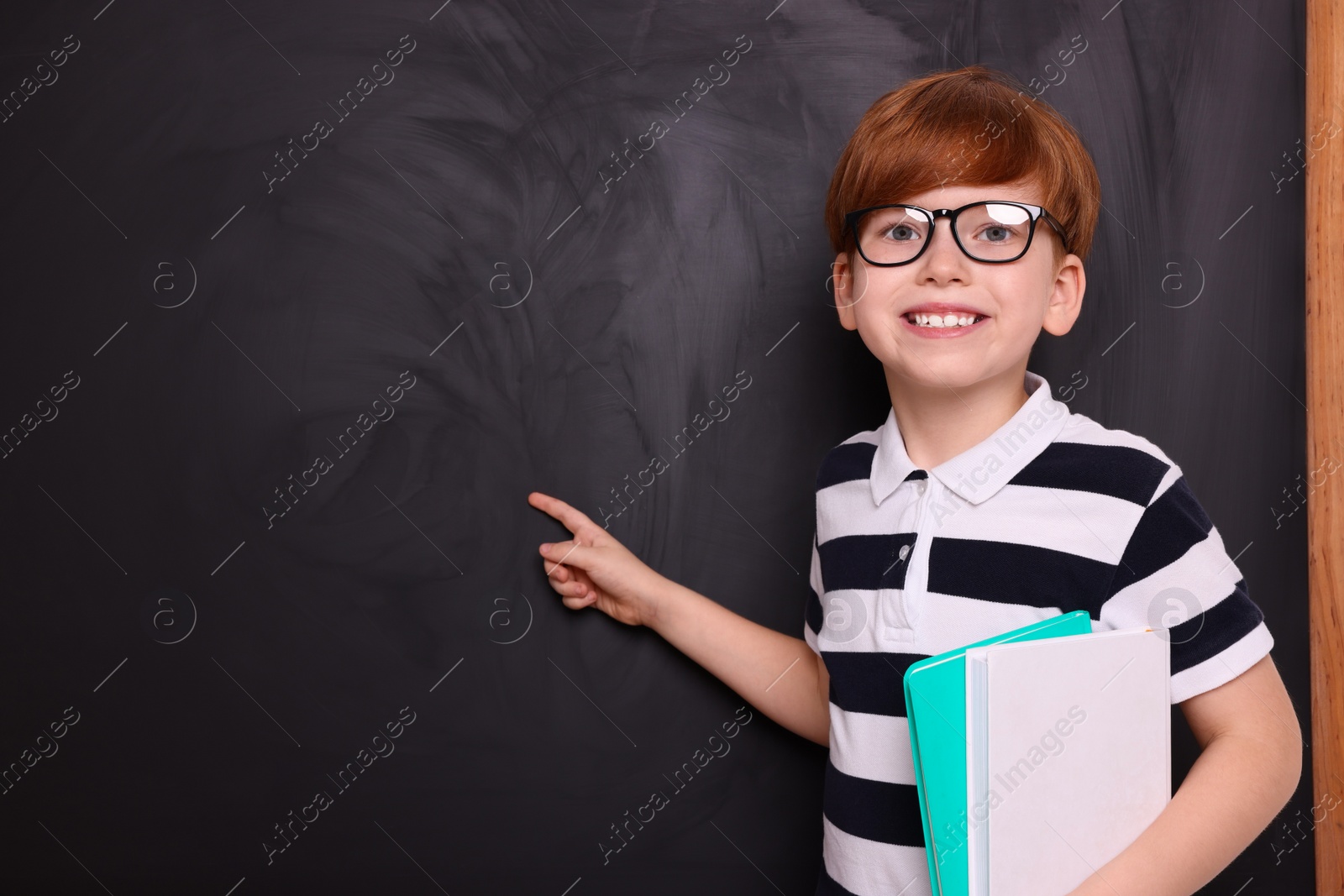 Photo of Smiling schoolboy in glasses with books pointing at something on blackboard. Space for text