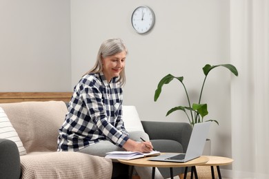 Photo of Beautiful senior woman writing something in notebook while using laptop at home