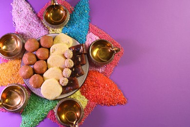 Photo of Diwali celebration. Tasty Indian sweets, diya lamps and colorful rangoli on violet table, flat lay. Space for text