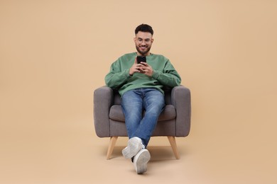 Happy young man using smartphone in armchair on beige background