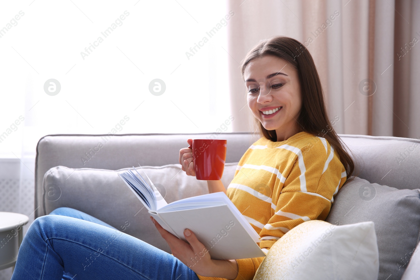 Photo of Young woman with cup of coffee reading book on couch at home