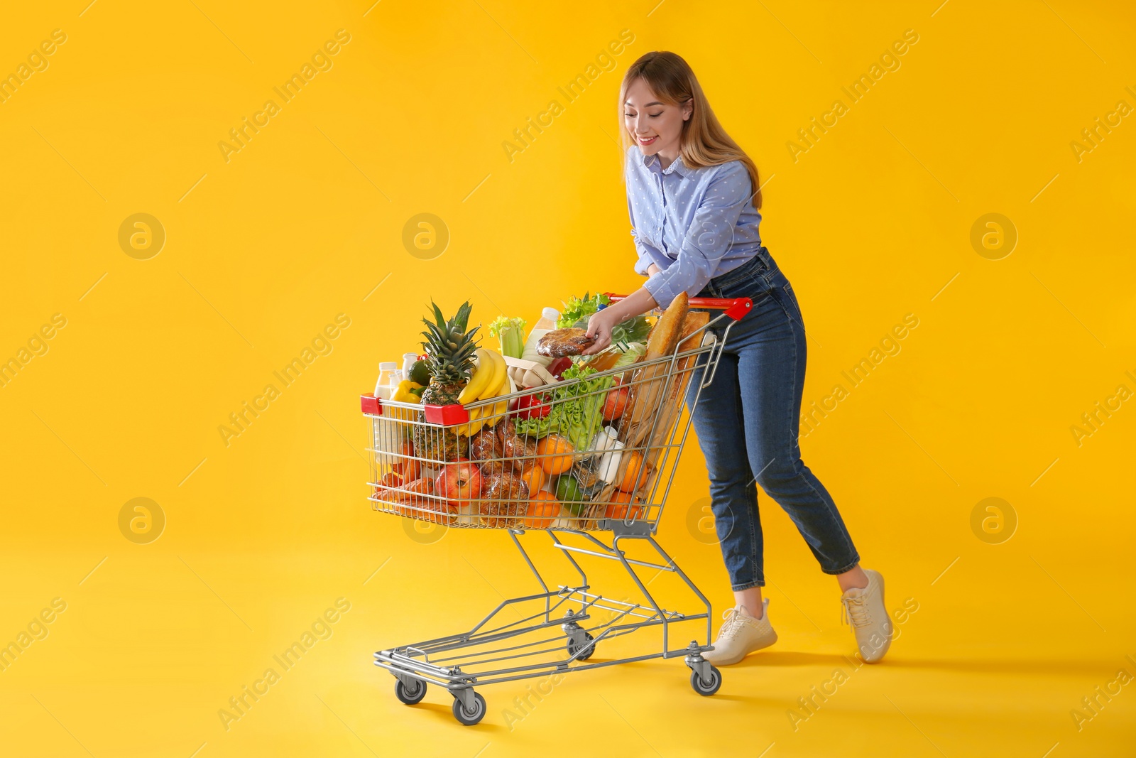 Photo of Young woman with shopping cart full of groceries on yellow background