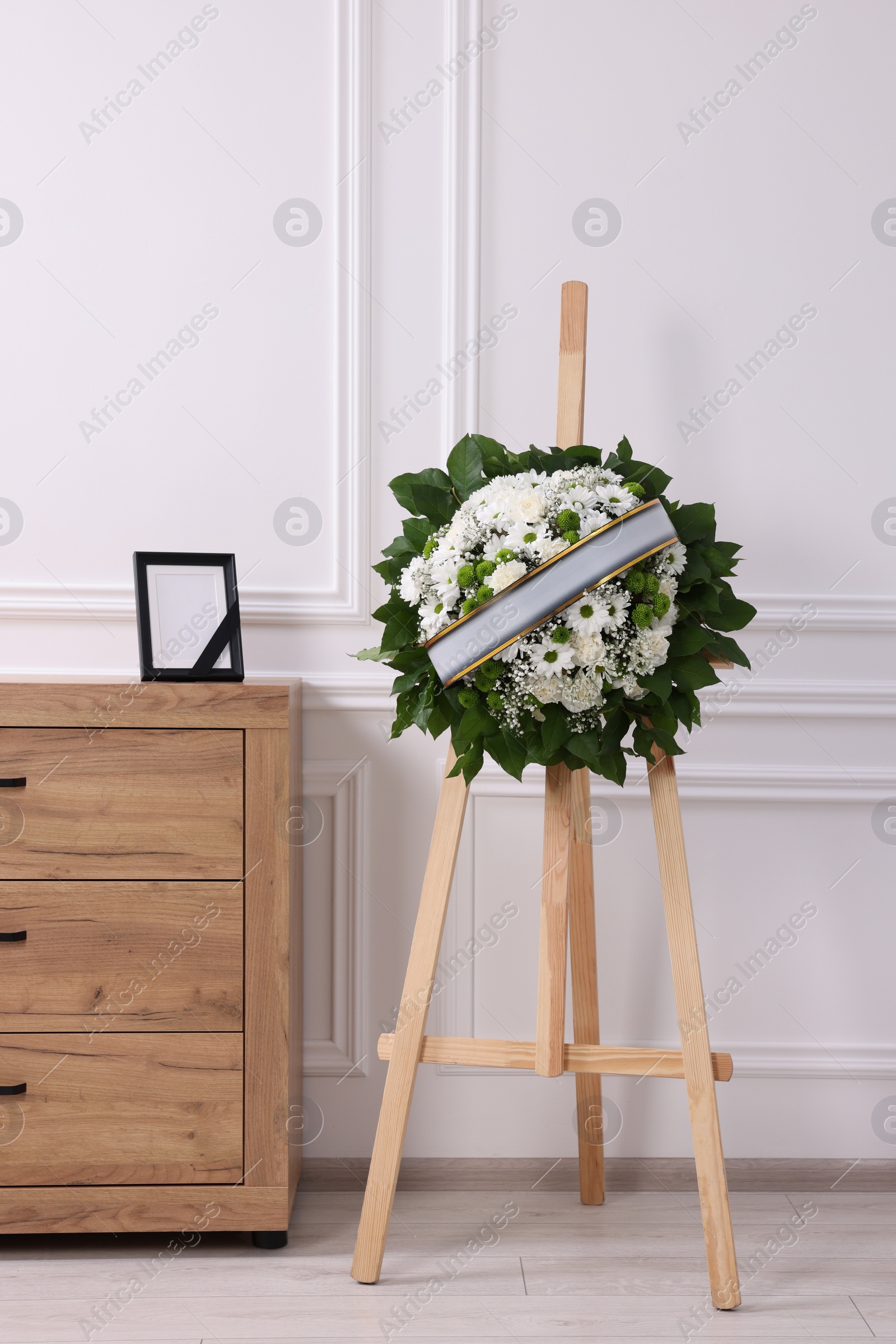 Photo of Wreath of flowers and photo frame with black ribbon on commode in room. Funeral attributes