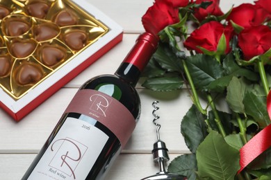 Bottle of red wine, beautiful roses and heart shaped chocolate candies on white wooden table, closeup