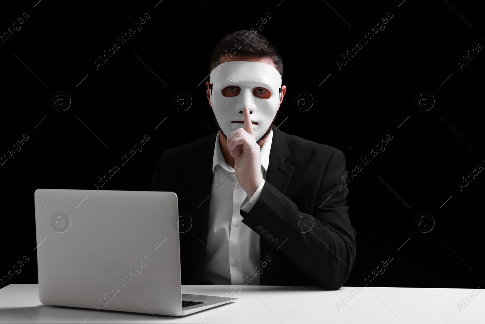 Photo of Man in mask and gloves sitting with laptop and showing hush gesture at white table against black background