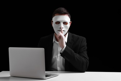 Man in mask and gloves sitting with laptop and showing hush gesture at white table against black background