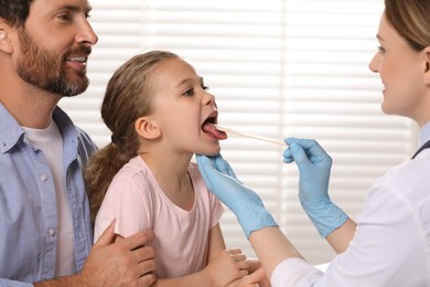 Photo of Doctor examining girl`s oral cavity with tongue depressor near her father indoors