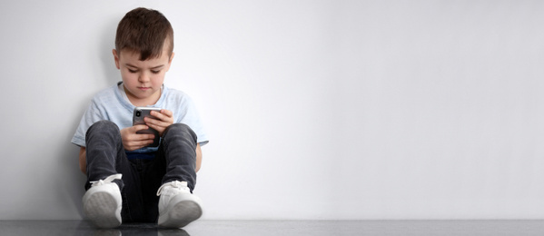 Image of Sad little boy with mobile phone sitting near white wall, banner design and space for text. Time to visit child psychologist