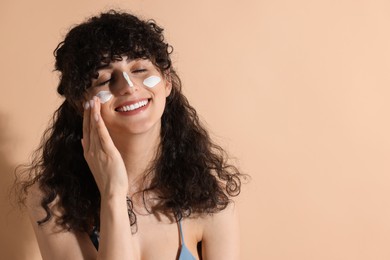 Beautiful young woman applying sun protection cream onto face on beige background, space for text