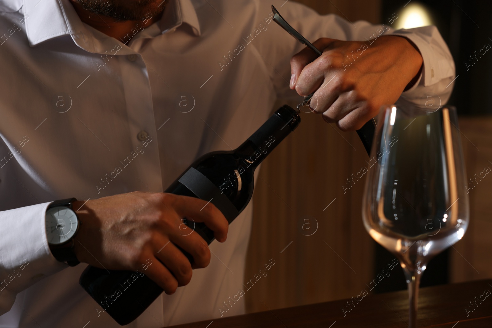 Photo of Romantic dinner. Man opening wine bottle with corkscrew at table indoors, closeup