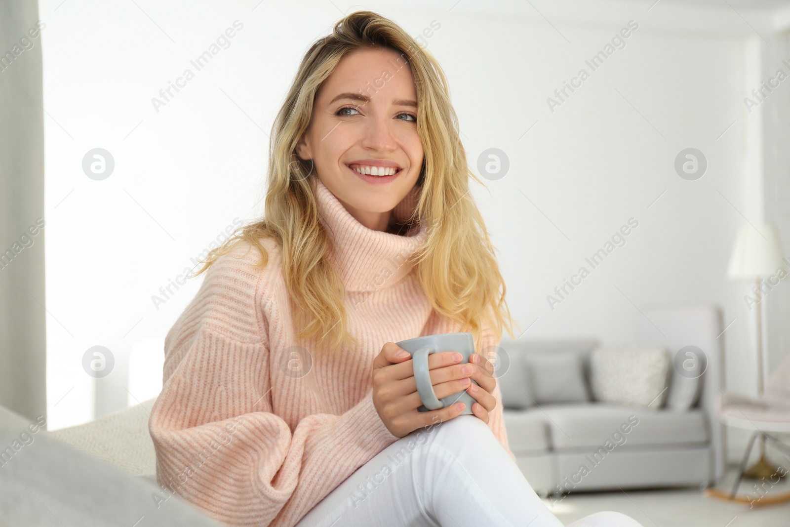 Photo of Beautiful young woman with cup of hot drink wearing warm pink sweater at home