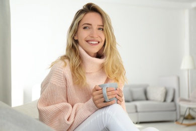 Photo of Beautiful young woman with cup of hot drink wearing warm pink sweater at home
