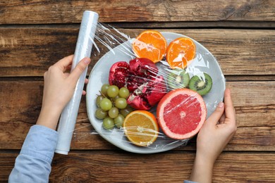 Photo of Woman putting plastic food wrap over plate of fresh fruits at wooden table, top view