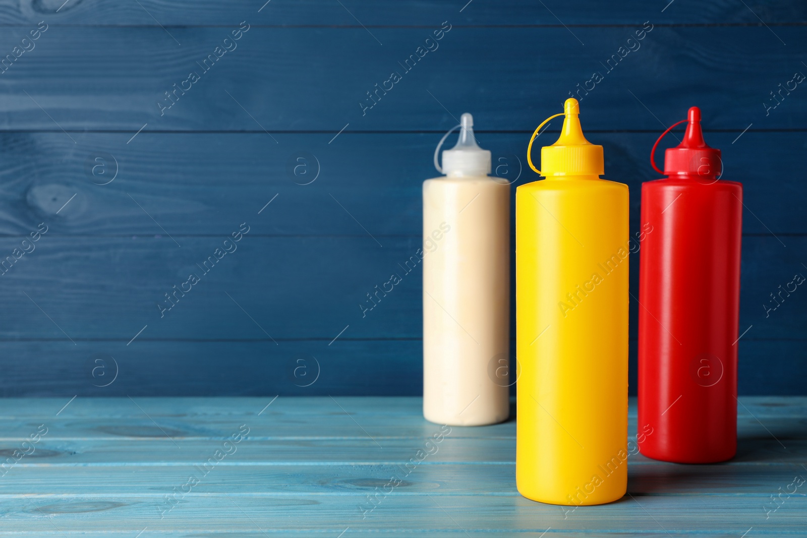 Photo of Bottles of mustard, ketchup and mayonnaise on blue wooden table. Space for text