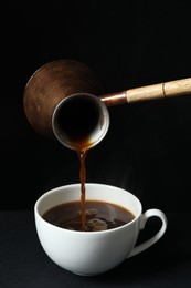 Photo of Turkish coffee. Pouring brewed beverage from cezve into cup on table against black background