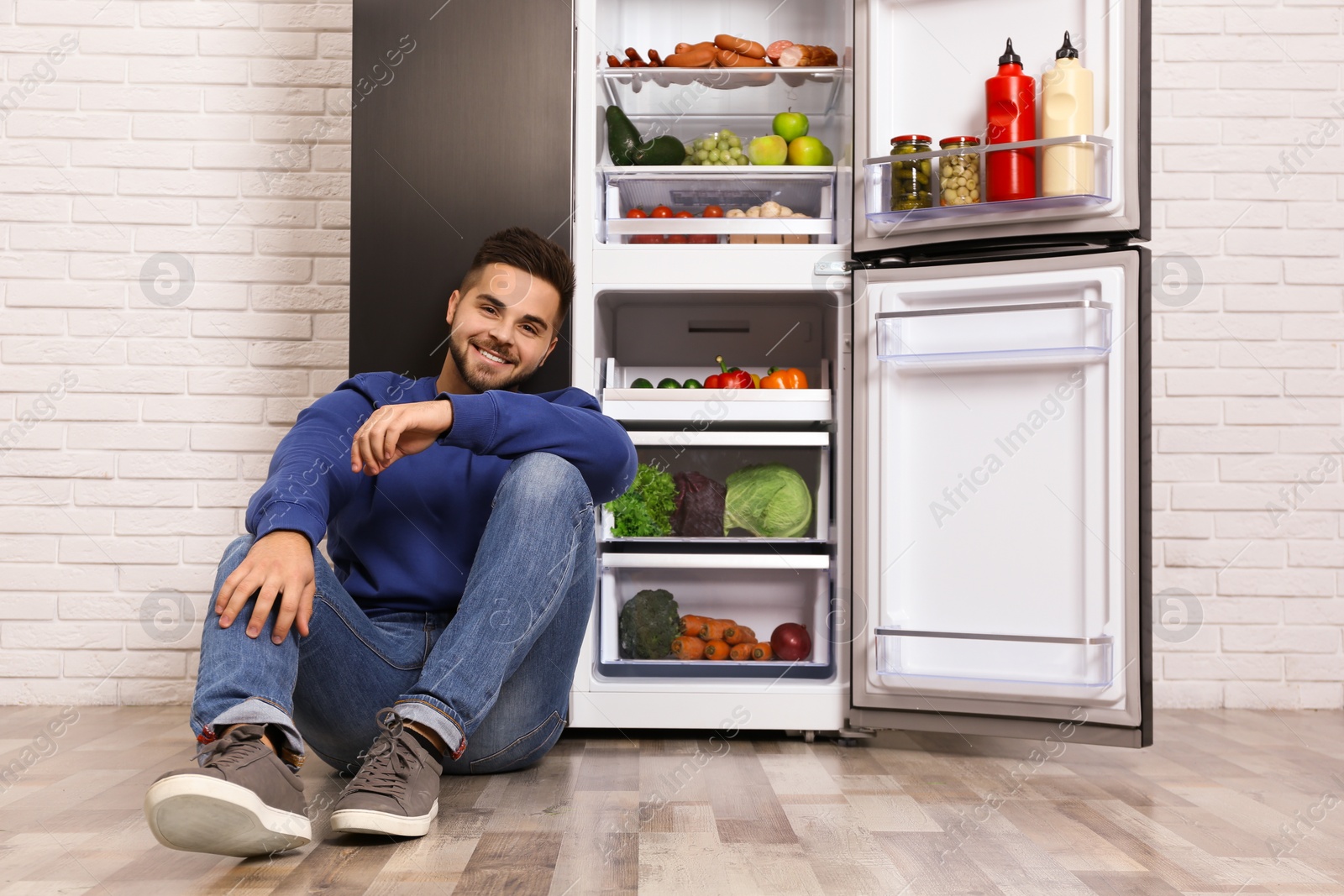 Photo of Happy young man sitting on floor near open refrigerator indoors