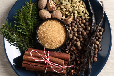 Plate with different aromatic spices and fir branches on light wooden table, top view