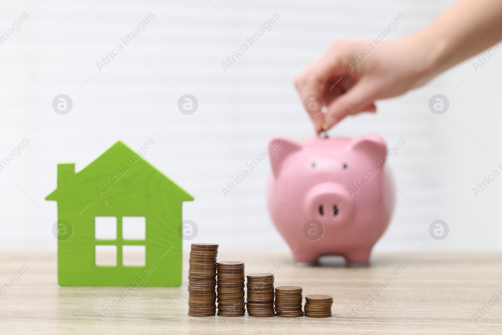 Photo of Woman putting coin into piggy bank at wooden table, focus on stacked coins