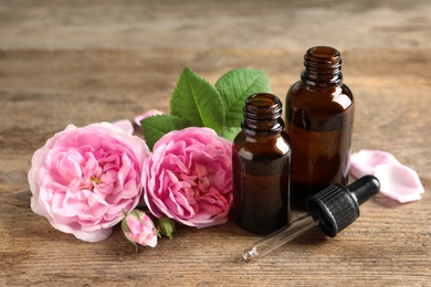 Photo of Bottles of rose essential oil, pipette and flowers on wooden table