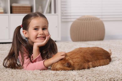 Happy little girl petting cute ginger cat on carpet at home