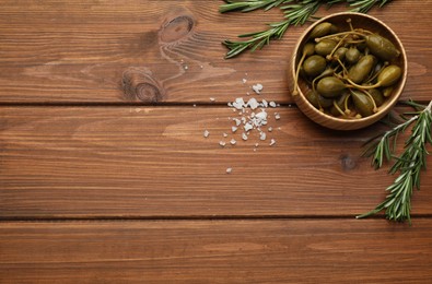 Photo of Tasty capers in bowl, salt and rosemary on wooden table, flat lay. Space for text