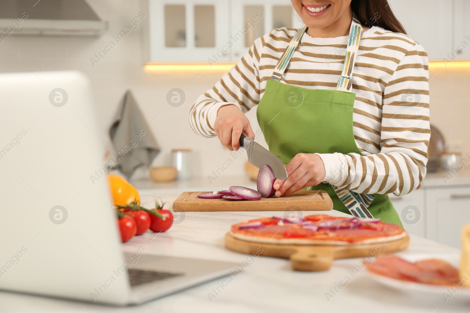 Photo of Happy woman cutting onion while watching online cooking course via laptop in kitchen, closeup