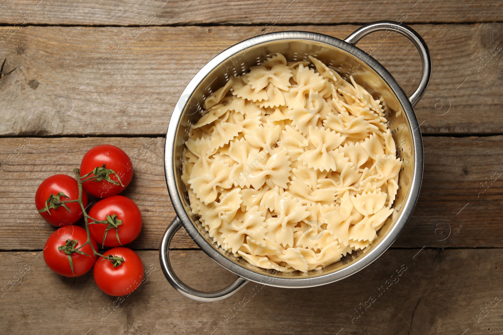 Photo of Cooked pasta in metal colander and tomatoes on wooden table, top view