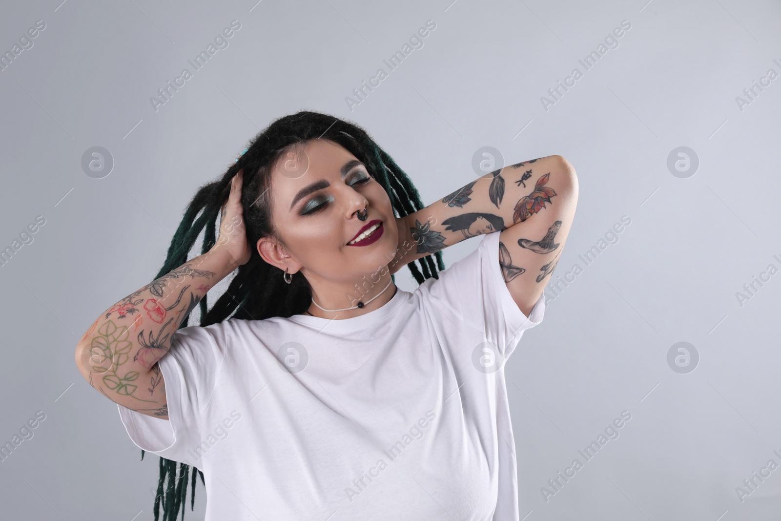 Photo of Beautiful young woman with tattoos on arms, nose piercing and dreadlocks against grey background