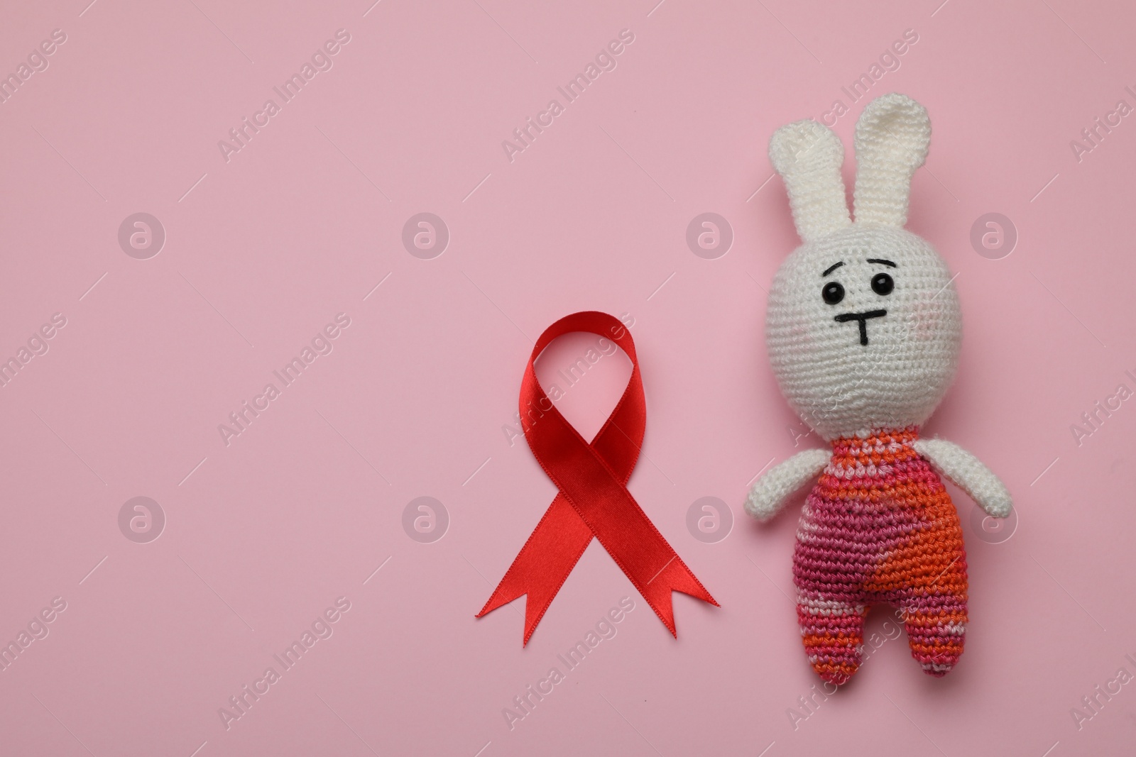 Photo of Cute knitted toy bunny and red ribbon on pink background, flat lay with space for text. AIDS disease awareness