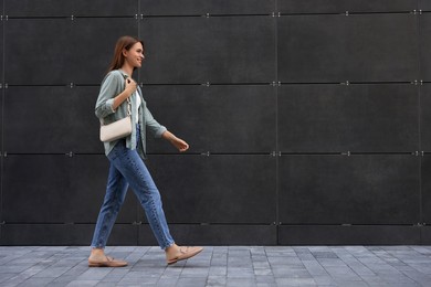 Young woman in casual clothes walking near grey wall outdoors, space for text
