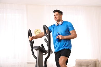 Photo of Man with bottle of water using modern elliptical machine at home