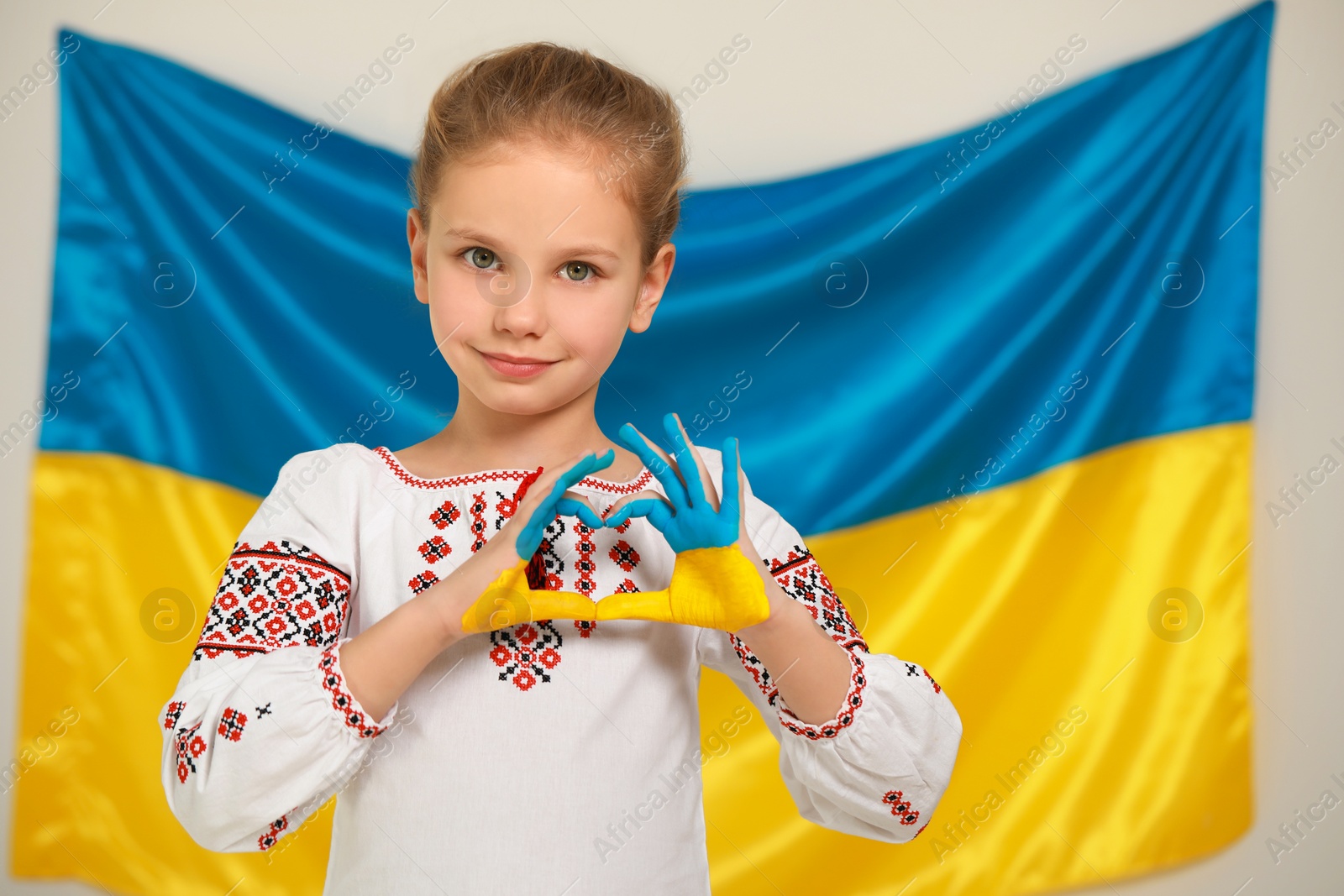 Photo of Little girl making heart with her painted hands near Ukrainian flag, space for text. Love Ukraine concept