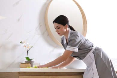 Photo of Young chambermaid wiping dust from countertop in bathroom