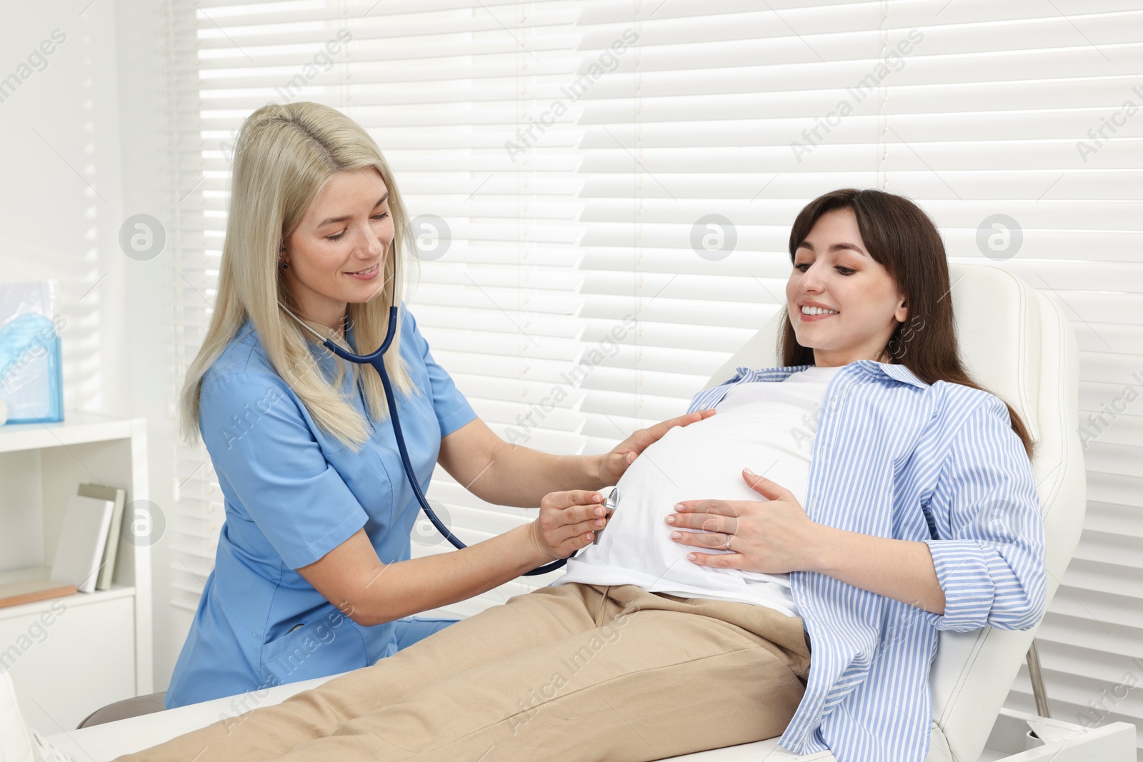 Photo of Pregnancy checkup. Smiling doctor with stethoscope listening baby's heartbeat in patient's tummy in clinic