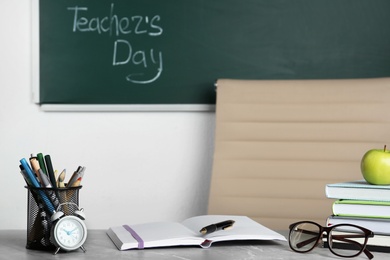 Photo of Eyeglasses and stationery on grey table near chalkboard with inscription TEACHER'S DAY in classroom