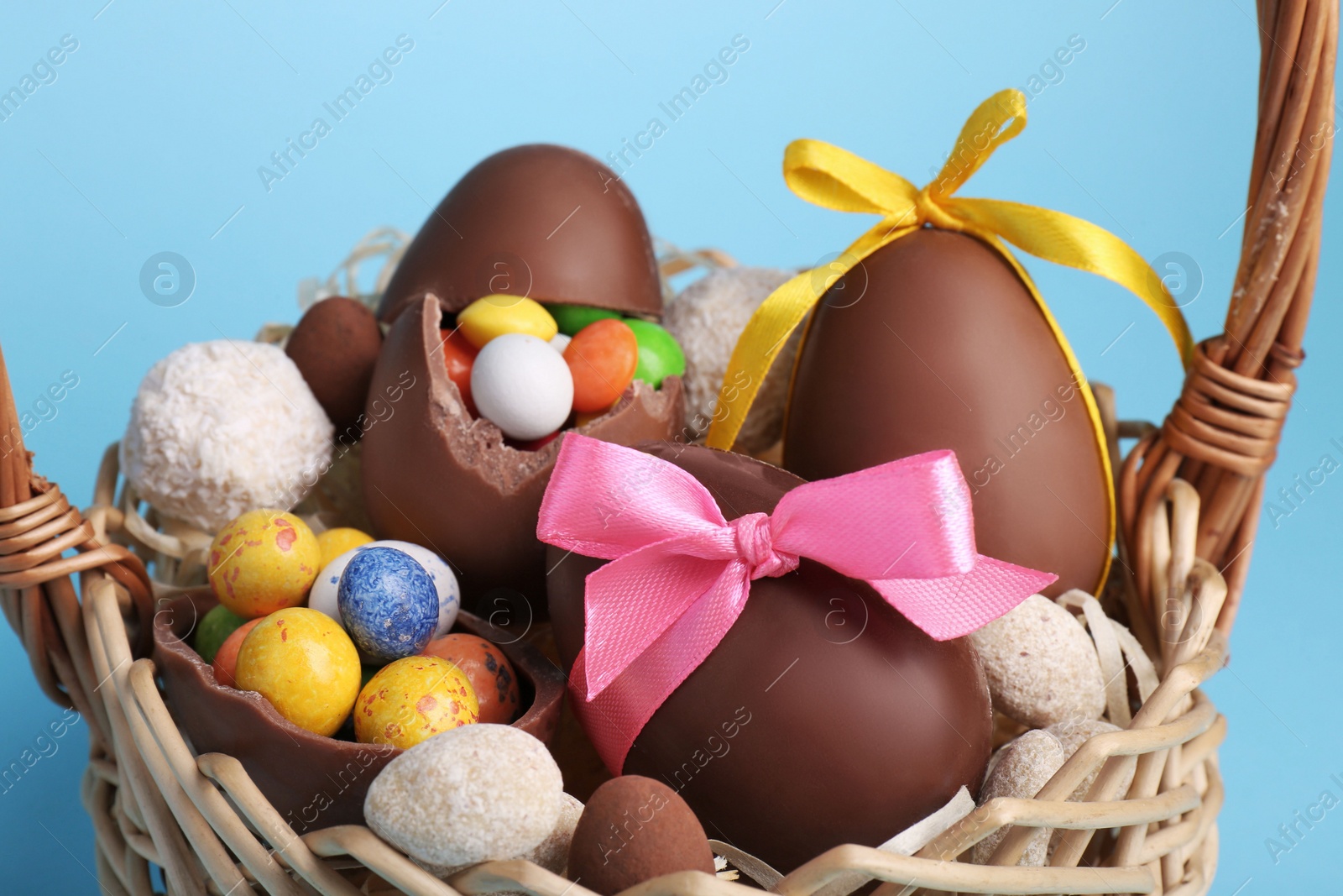 Photo of Wicker basket with tasty chocolate Easter eggs and different candies on light blue background, closeup