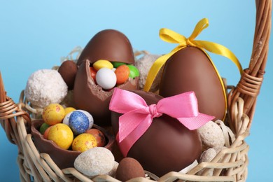 Wicker basket with tasty chocolate Easter eggs and different candies on light blue background, closeup