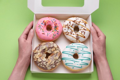 Photo of Woman holding box with tasty glazed donuts on light green background, top view