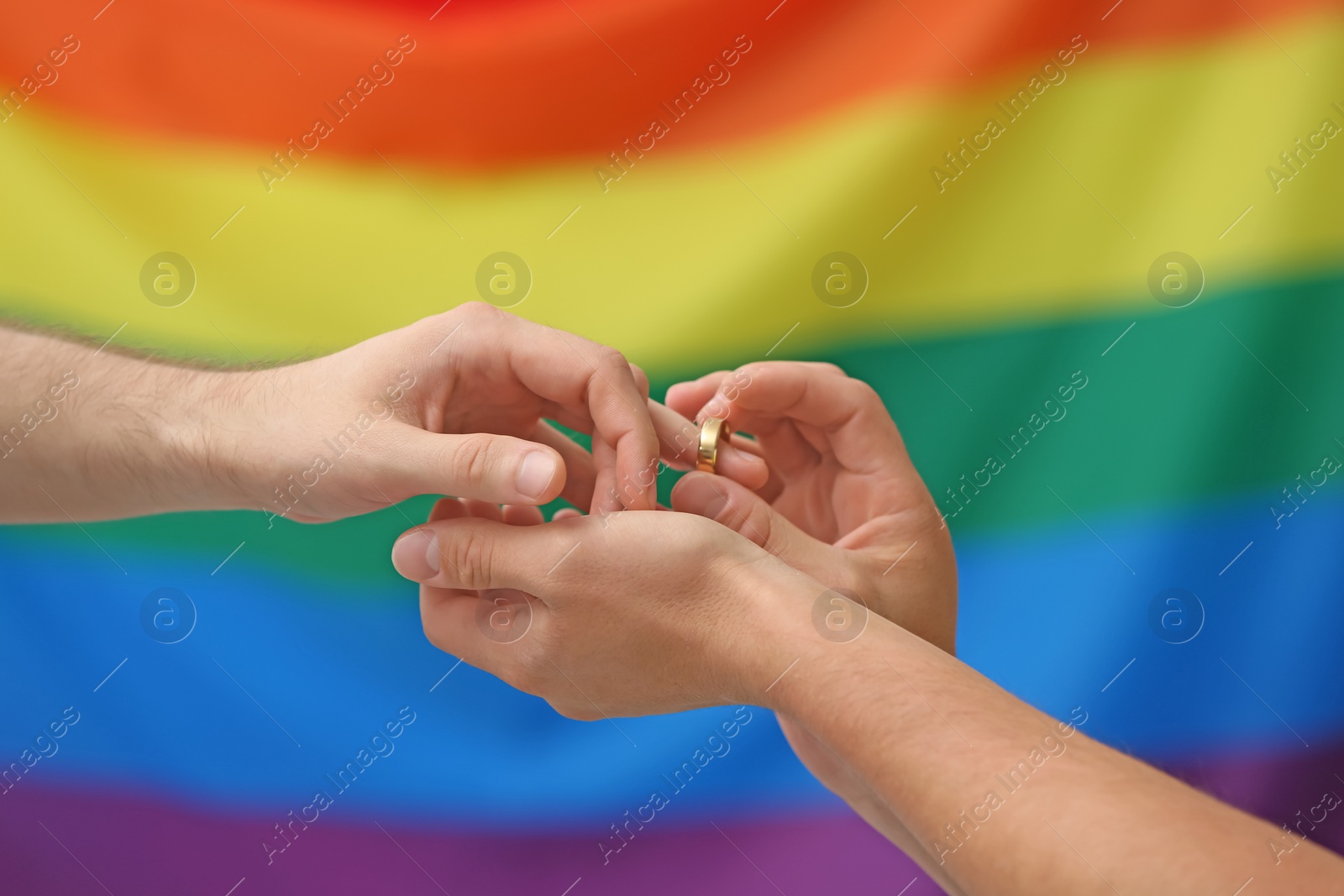 Photo of Young man putting wedding ring on his boyfriend's finger against rainbow background. Gay marriage