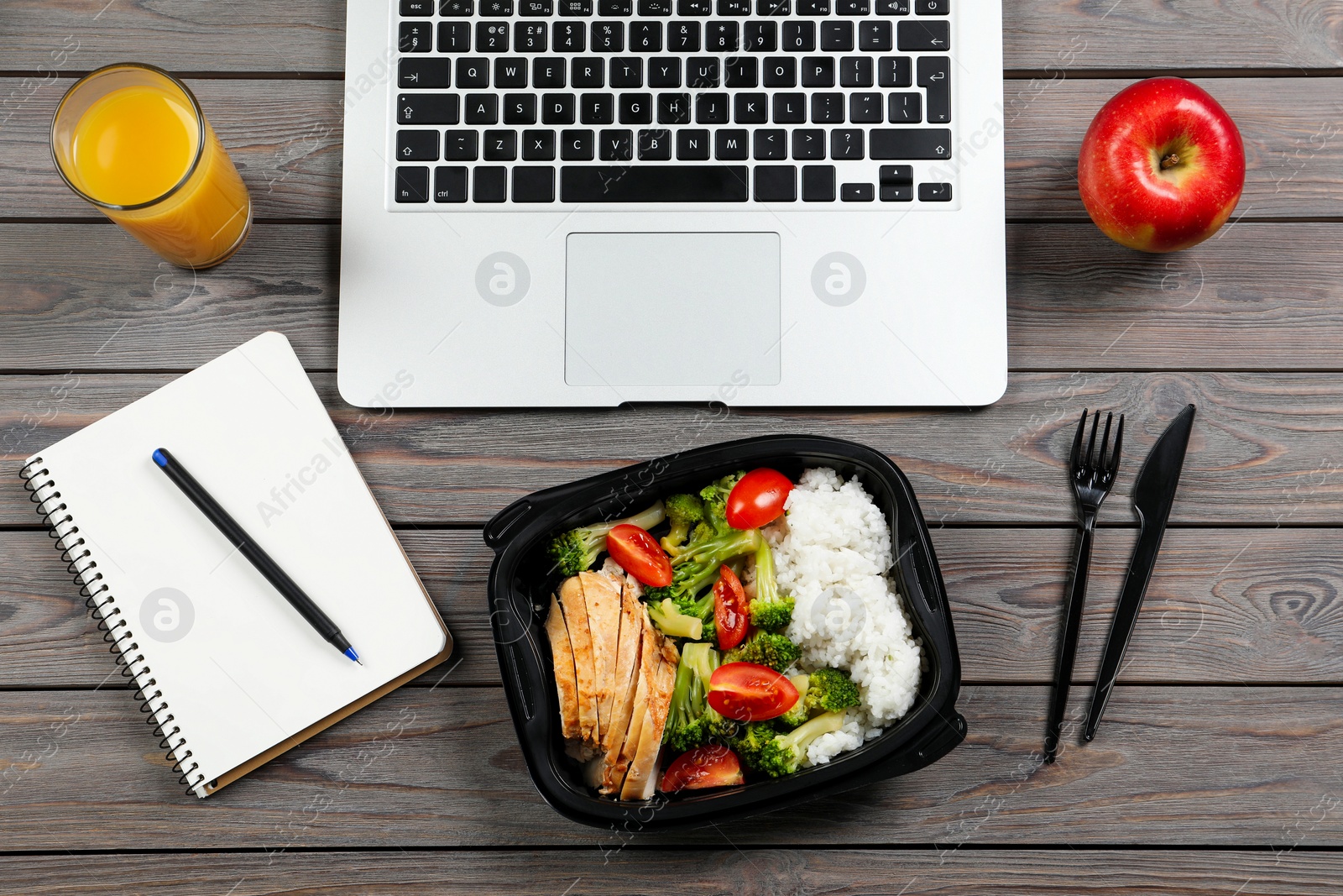 Photo of Container with tasty food, glass of juice, laptop and apple on wooden table, flat lay. Business lunch