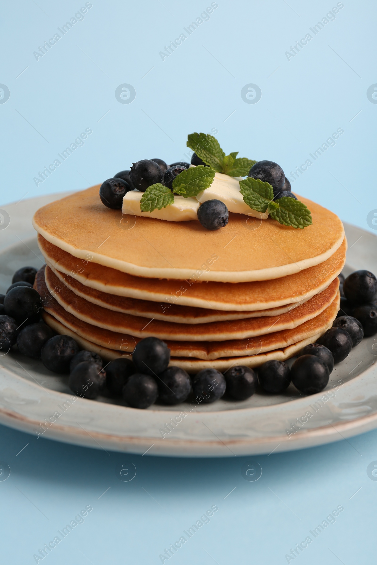 Photo of Stack of tasty pancakes with blueberries, butter and mint on light blue background, closeup