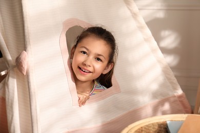Cute little girl peeking out window of toy wigwam at home