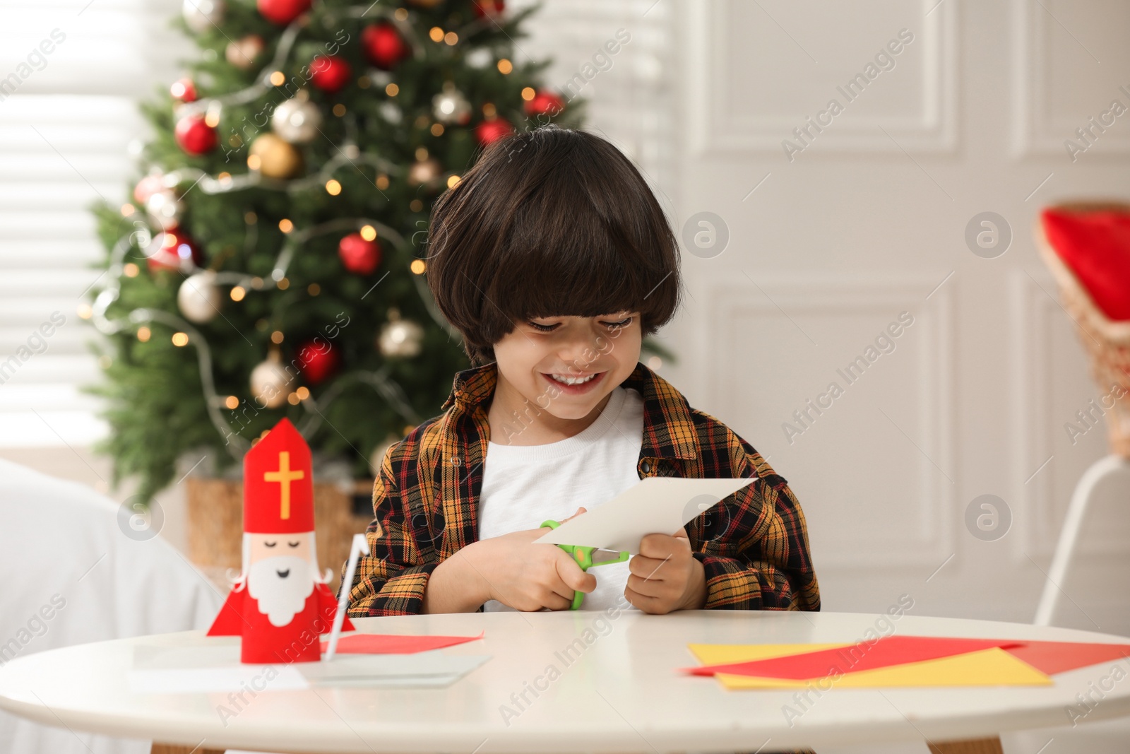 Photo of Cute little boy cutting paper at table with Saint Nicholas toy indoors